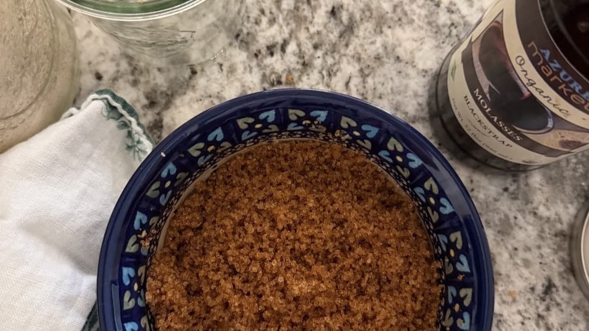 Overhead view of homemade brown sugar in a blue patterned bowl beside a jar of white sugar, a folded kitchen towel, and a bottle of blackstrap molasses.